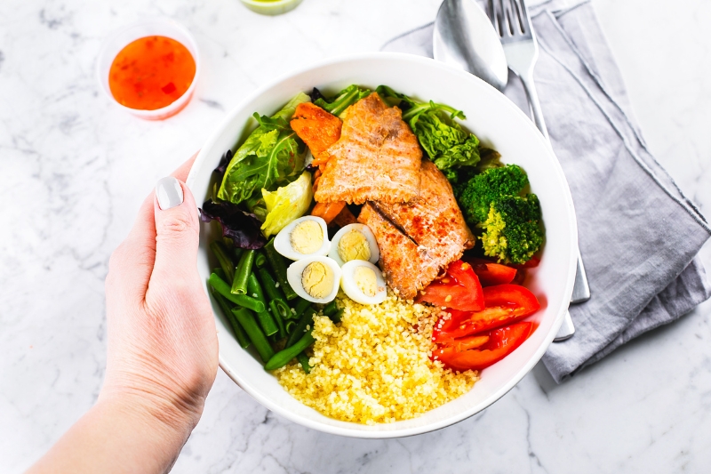 A person presents a bowl of grilled salmon, quinoa, and roasted garlic broccoli, highlighting a healthy dinner option.