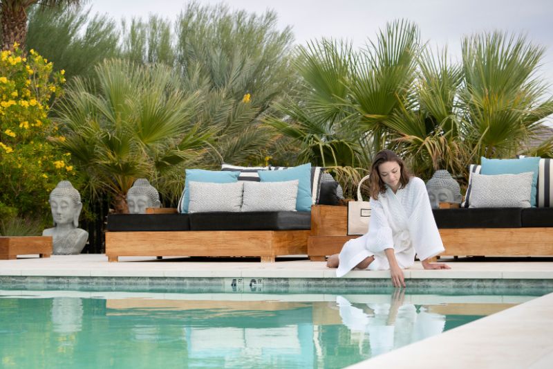 A person in a white robe sitting by a serene pool, gently touching the water, surrounded by lush greenery and comfortable lounge seating, evoking a peaceful and rejuvenating atmosphere.