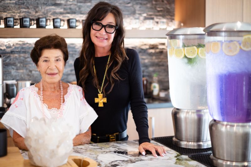 Two women standing together in a kitchen, one older and one younger, smiling while surrounded by infused water dispensers with lemon slices.