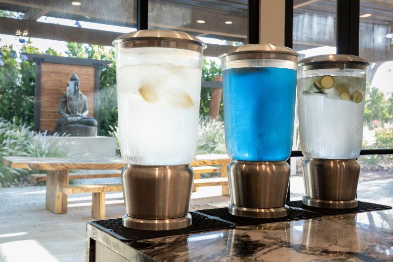 Three dispensers filled with colorful juices—lemon, cucumber, and a blue beverage—placed on a counter with a serene outdoor view featuring a Buddha statue.