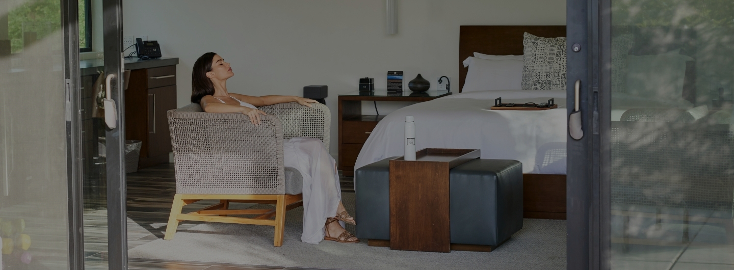 A woman seated on a chair in a luxurious wellness retreat, with a bed visible in the background.