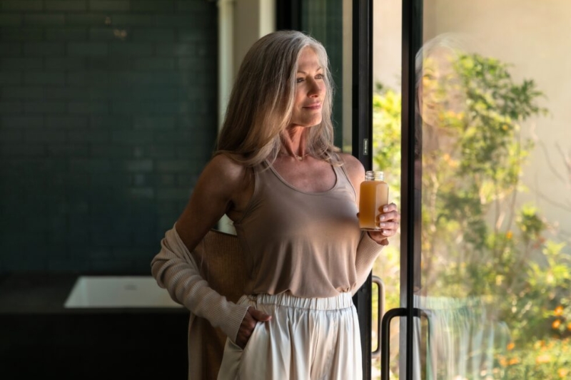 A woman stands by a window holding a glass of juice, symbolizing the benefits of a juice cleanse for gut health.