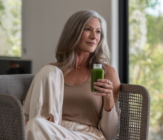 A woman with gray hair sits comfortably on a couch, sipping a green smoothie related to her Holistic Detox Program.