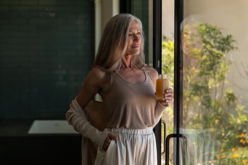 A woman stands by a window holding a glass of orange juice, promoting a Liquid Cleanse Detox lifestyle.