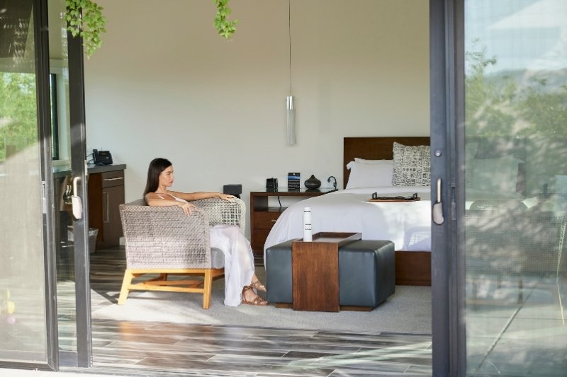A woman relaxes in a chair by a sliding glass door, with sunlight streaming into the room, creating a serene atmosphere.