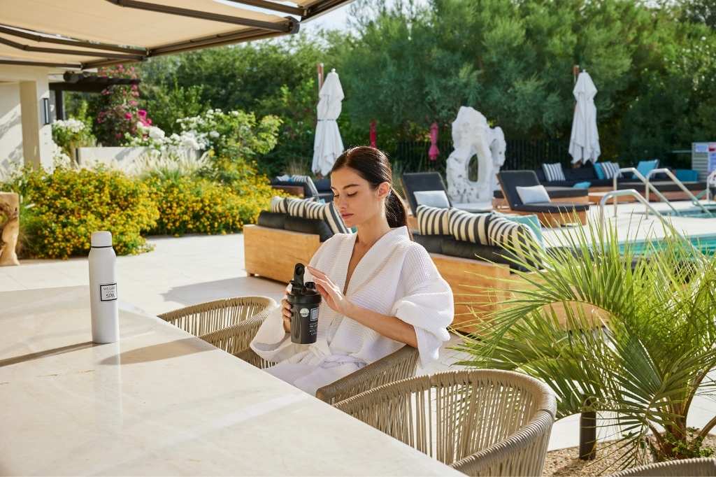A woman wearing a white robe relaxes at a table with a bottle of water, set in a Colon Hydrotherapy Spa environment.
