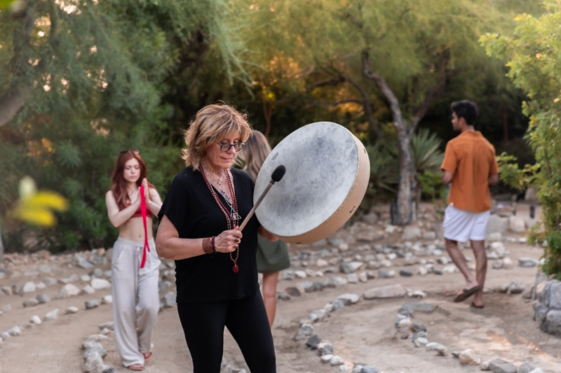A woman engaged in drumming within a circle of rocks, reflecting a holistic philosophy of wellness and detox support.