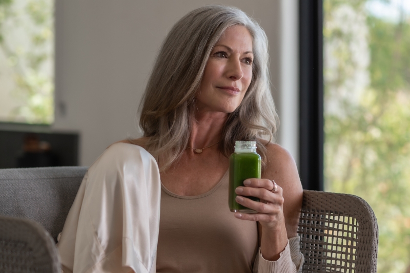 A gray-haired woman holds a bottle of green juice by a window.
